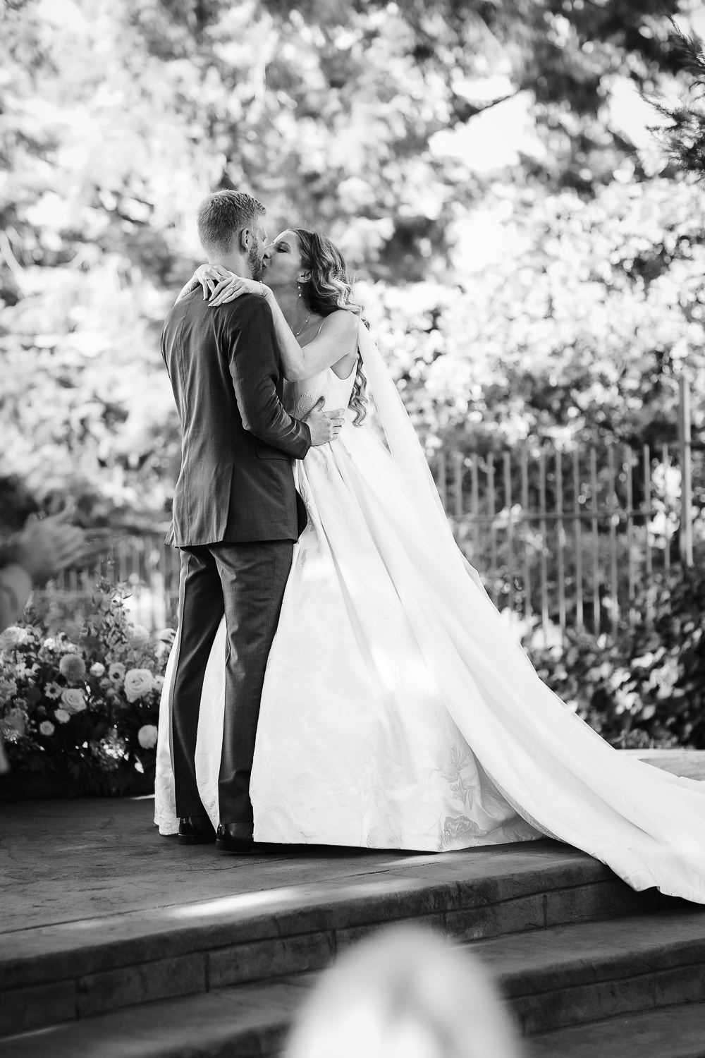 A romantic black and white wedding photo showing a couple embracing on an outdoor terrace surrounded by lush foliage.