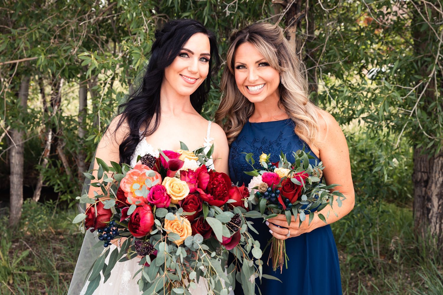 Two people holding colorful red and peach floral bouquets pose outdoors in formal dresses against a natural background.