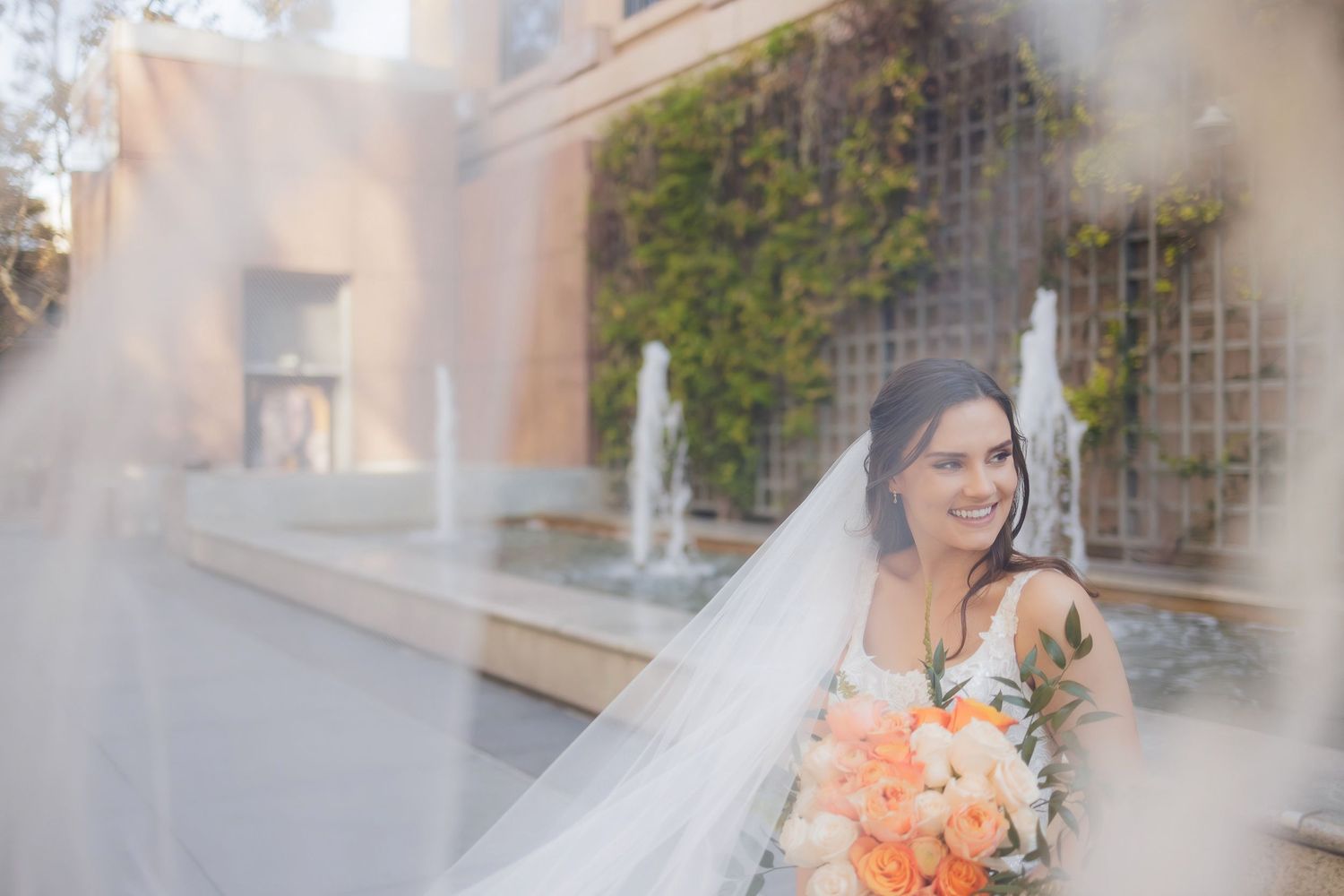 Elegant bride holding coral bouquet near outdoor fountain with veil flowing in soft focus.