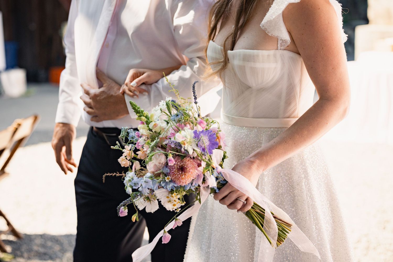 A close-up view of a bridal bouquet with delicate purple and white flowers against a white dress and dark pants.