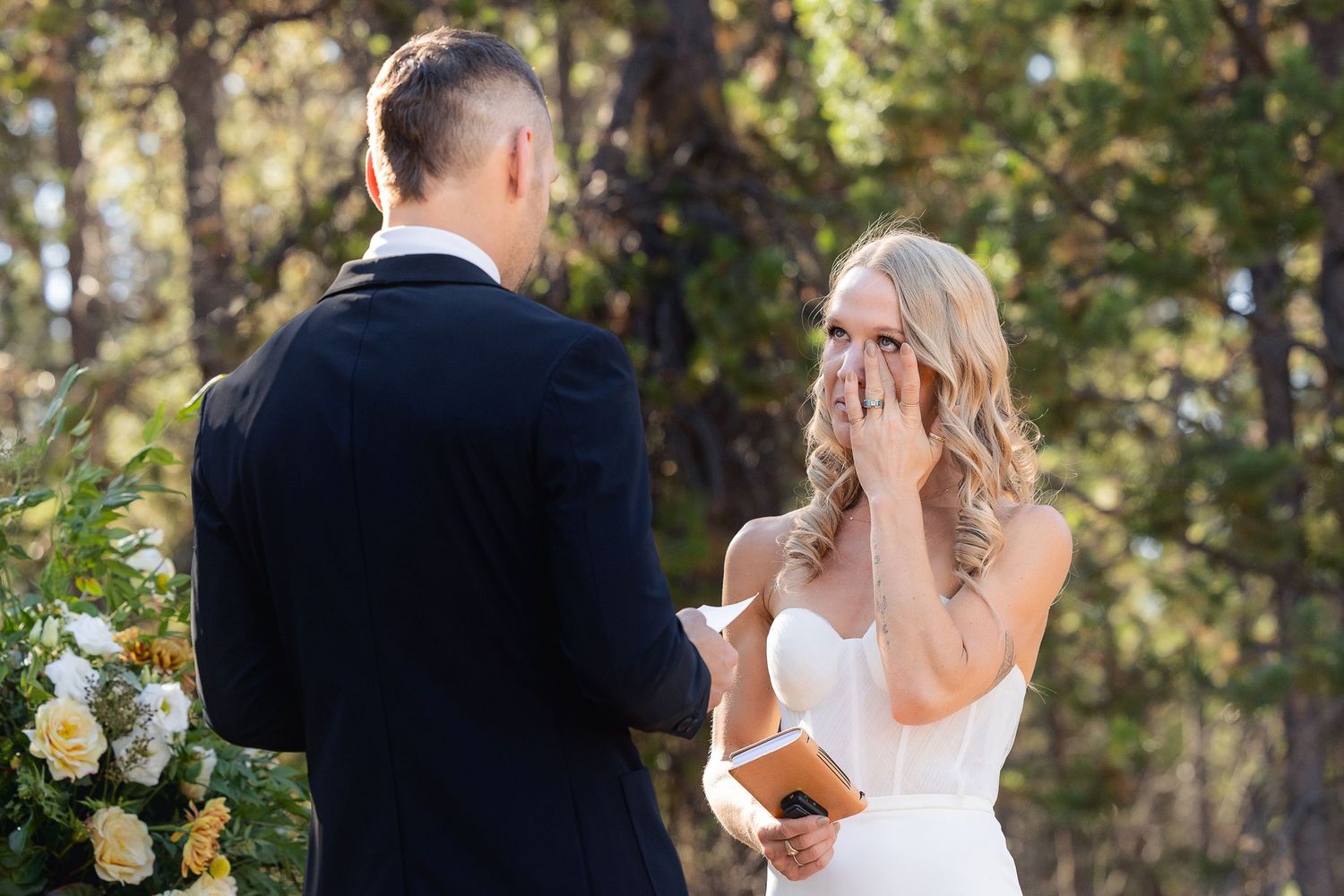 Bride reacts with emotion during outdoor forest wedding ceremony with floral arrangements and natural backdrop.