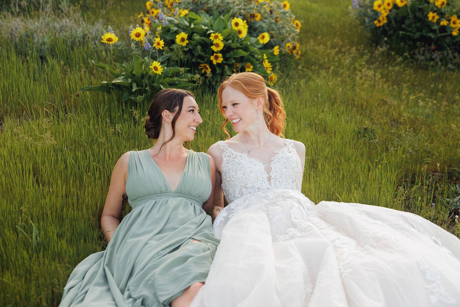 Bride in white gown and bridesmaid in sage green dress sitting together in a sunflower field.