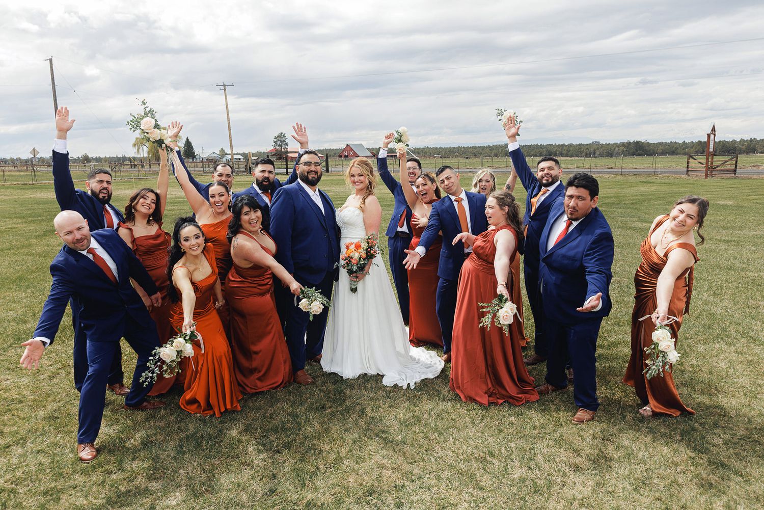 Wedding party in navy suits and rust bridesmaid dresses celebrating outdoors with bride and groom on a cloudy day.