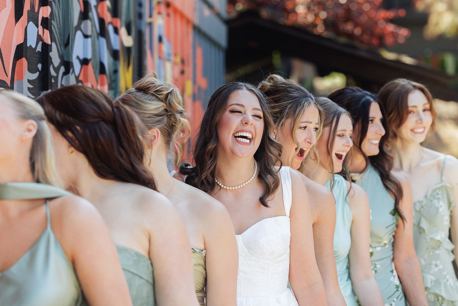 Bride in white gown laughing joyfully with bridesmaids in sage green dresses outdoors.