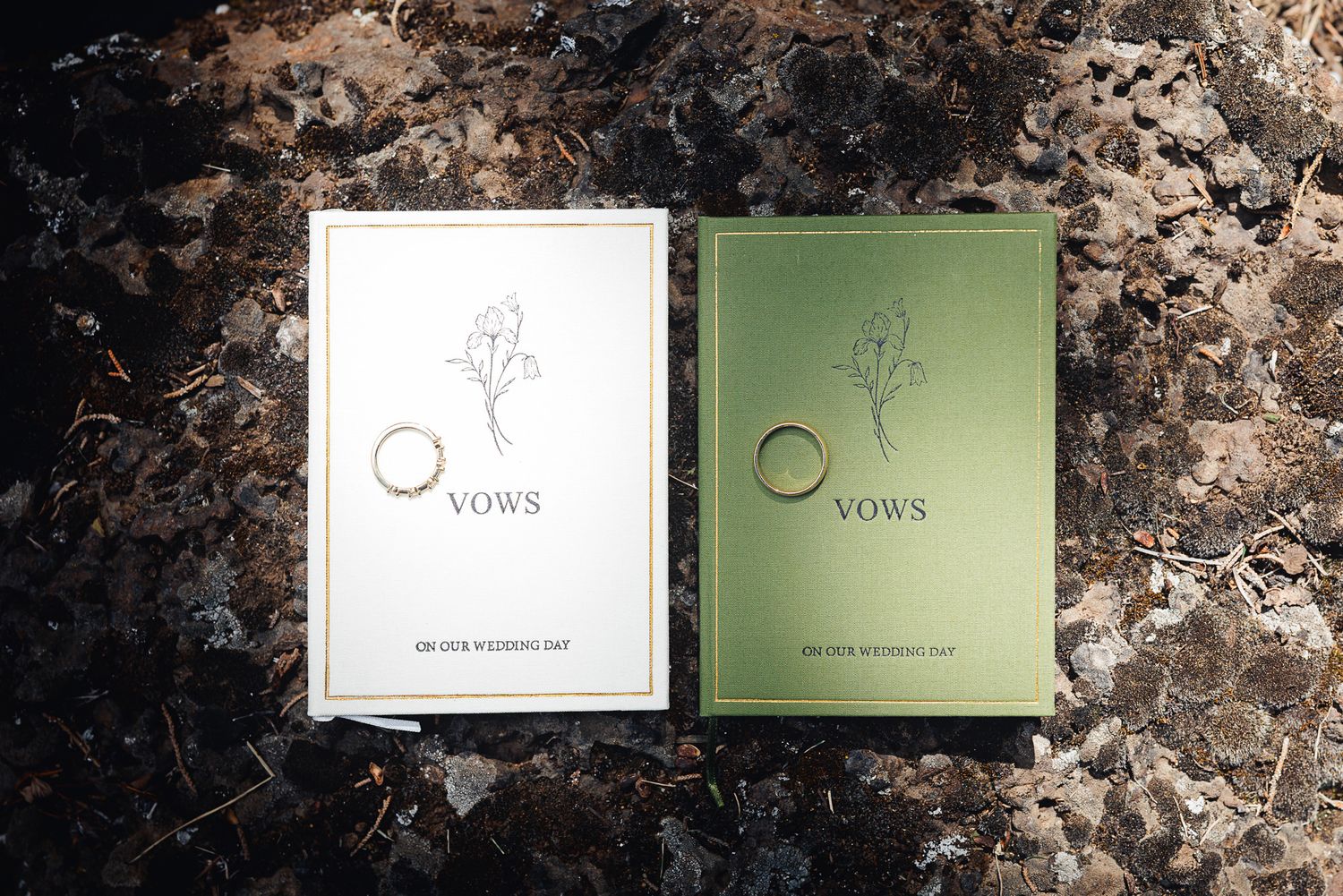 Two elegant wedding vow books, one white and one green, with gold rings resting on top, placed on a dark textured surface.