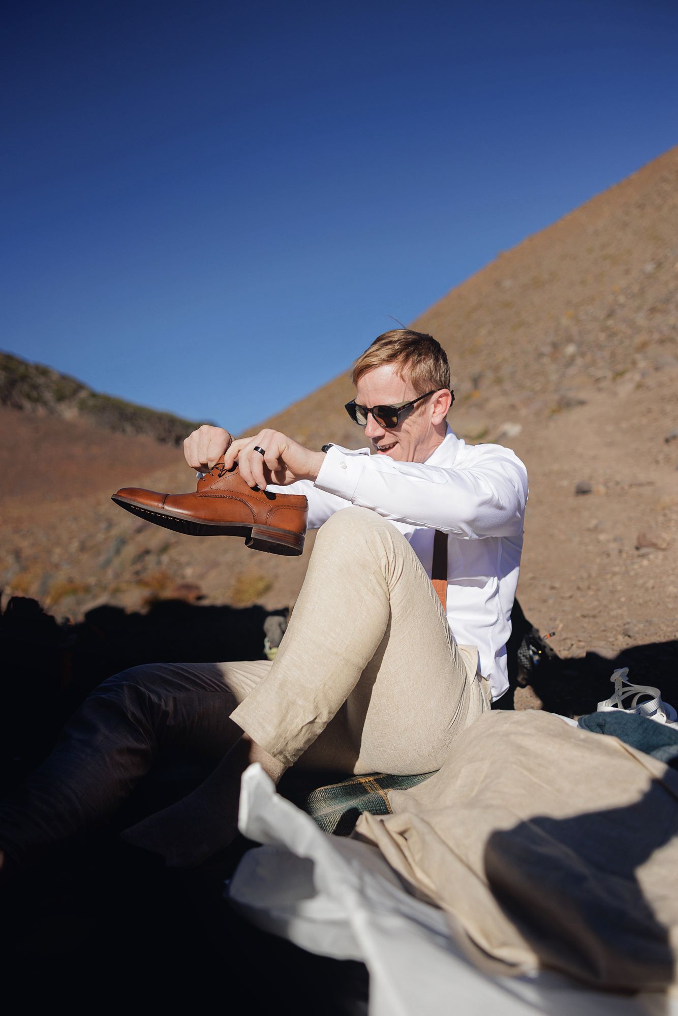 Man in sunglasses and a white shirt sits on a blanket in a desert landscape, holding a brown leather dress shoe under a clear blue sky.