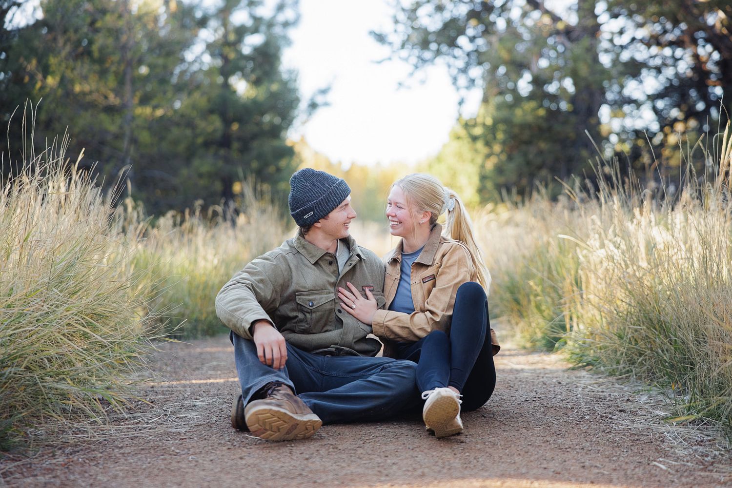 A smiling couple in casual jackets sits close together on a dirt path, laughing and relaxing among tall golden grass and trees.