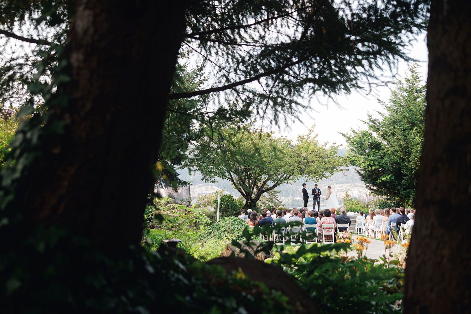 Outdoor wedding ceremony viewed through lush green trees with guests gathered near a waterfront venue.