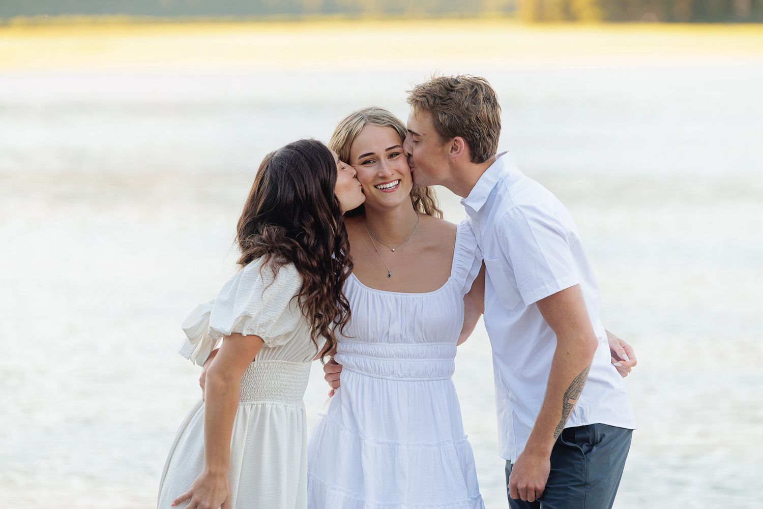 A young woman in white dress smiles as two people kiss her cheeks near a sunlit lake.