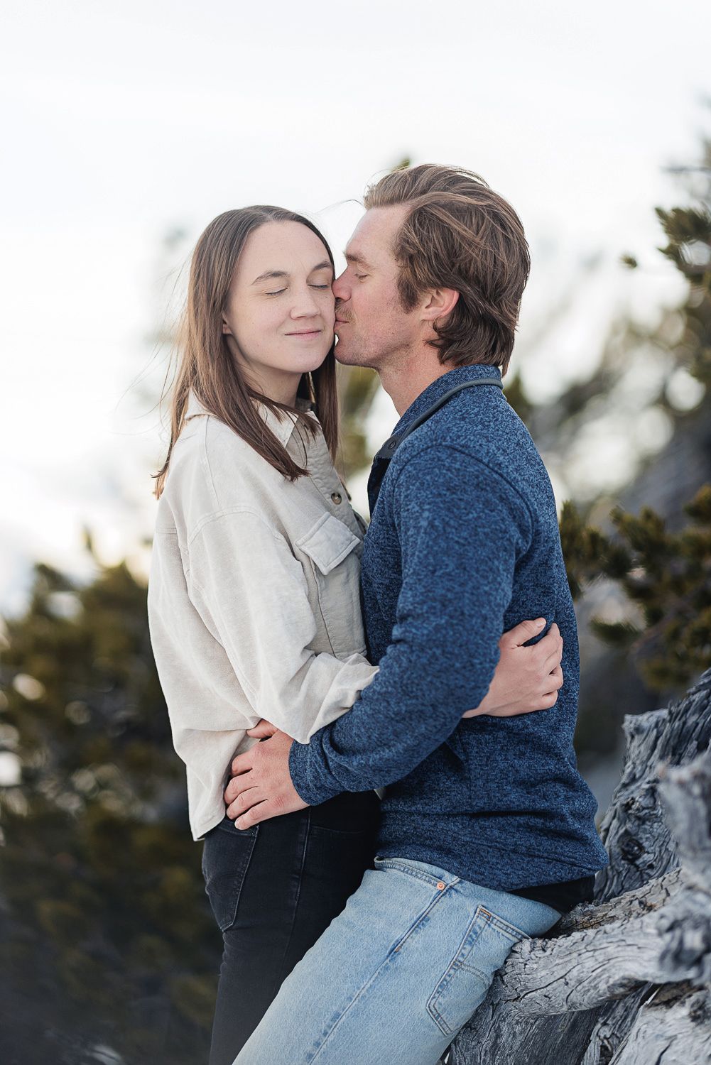 A couple in a blue sweater and white jacket embrace while sitting on a rock outdoors during a winter photo session.