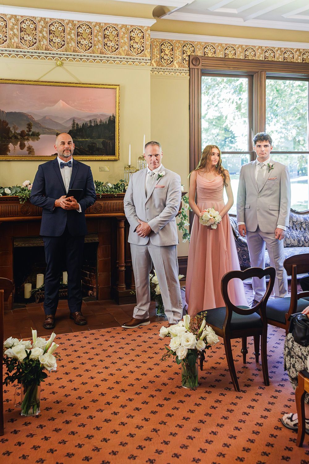 Four people pose playfully on a carpeted floor with ornate patterned wallpaper and vintage decor in the background.