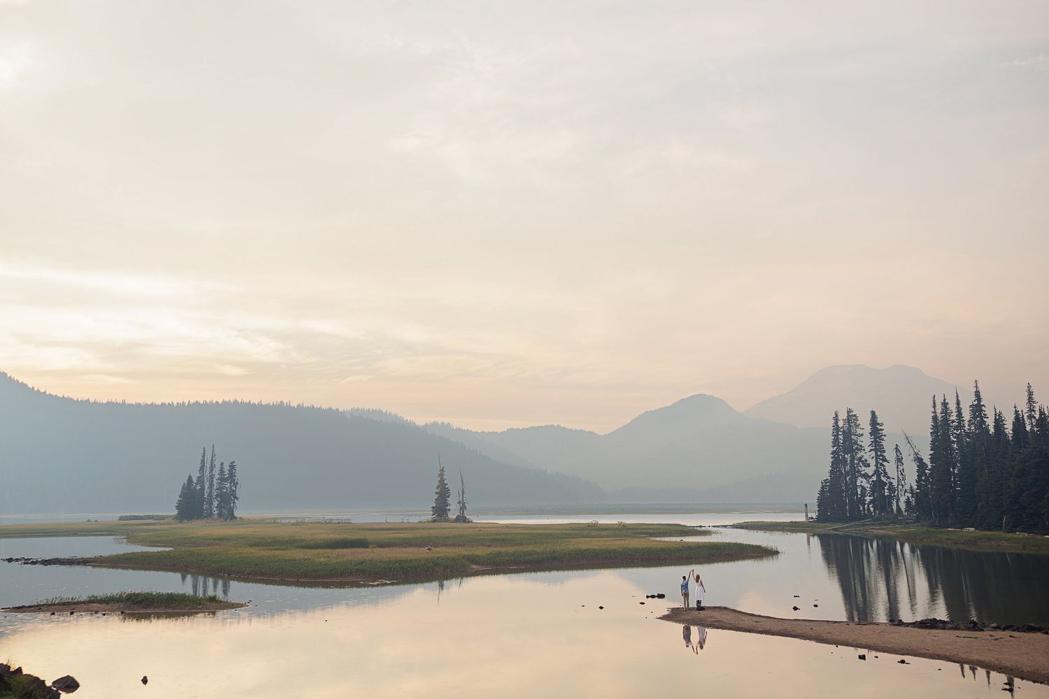 A tranquil mountain lake surrounded by evergreen trees and misty peaks reflects golden sunset light in serene waters.