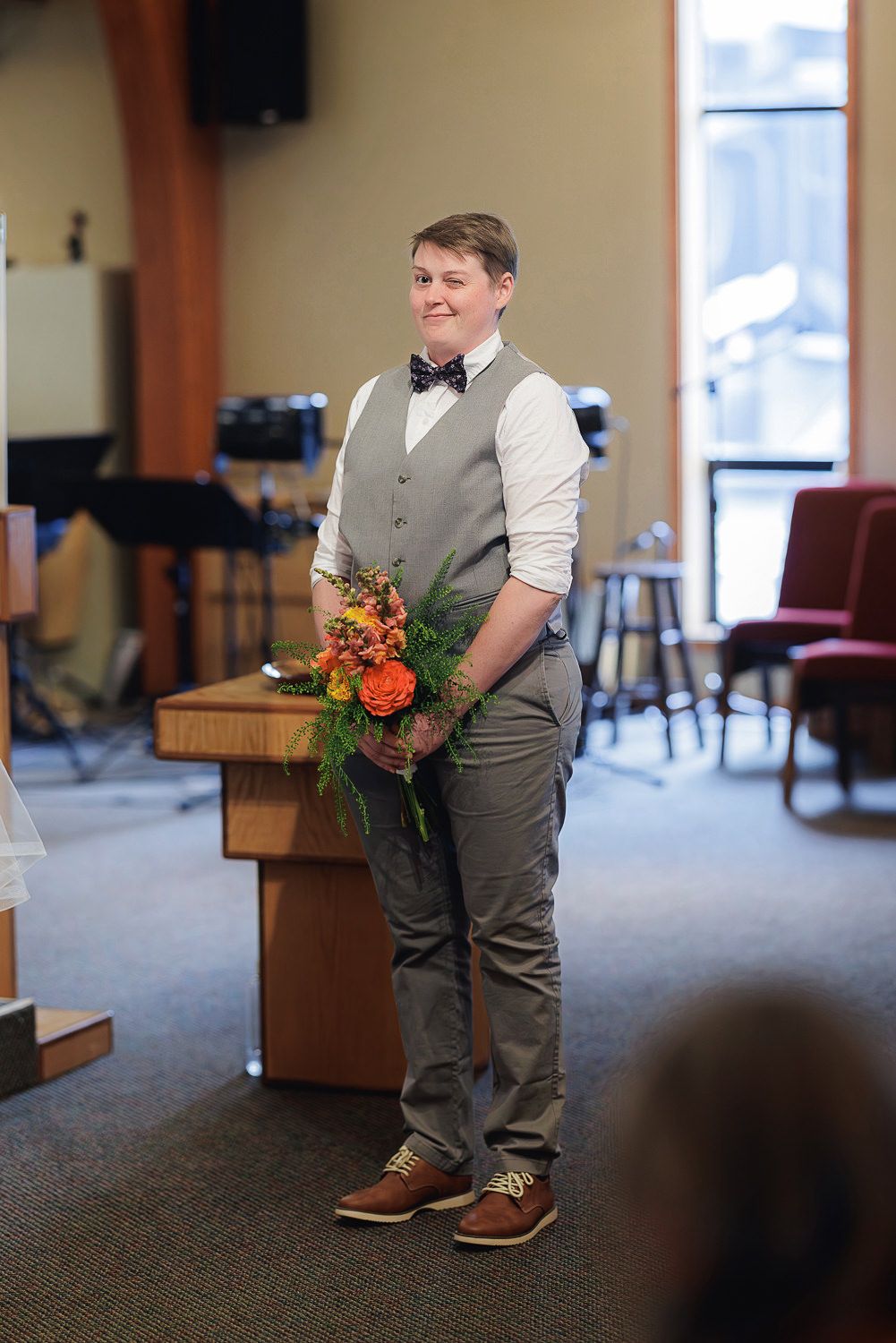 Groomsman in gray vest and khaki pants stands in a carpeted room holding orange and pink floral bouquet.