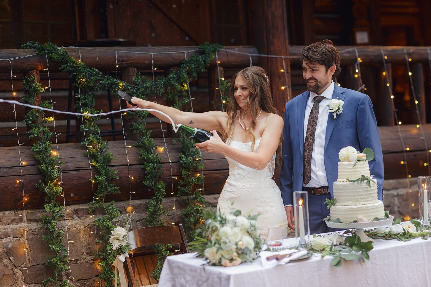 A couple cuts their white wedding cake together during an indoor reception with string lights and greenery decorations.