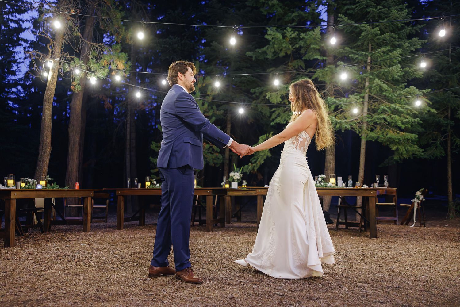 A couple dances together under twinkling lights strung between trees in a romantic outdoor evening setting.