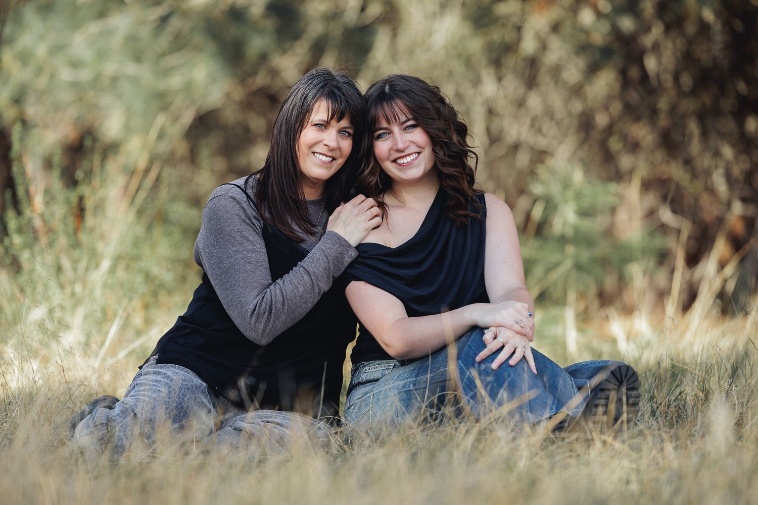 Two smiling women with dark hair sit together in a grassy outdoor field surrounded by trees.