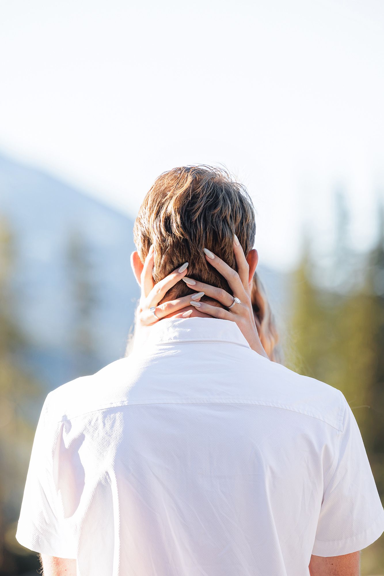 A woman's hand with an engagement ring rests on a man's neck outdoors with mountains in the background.