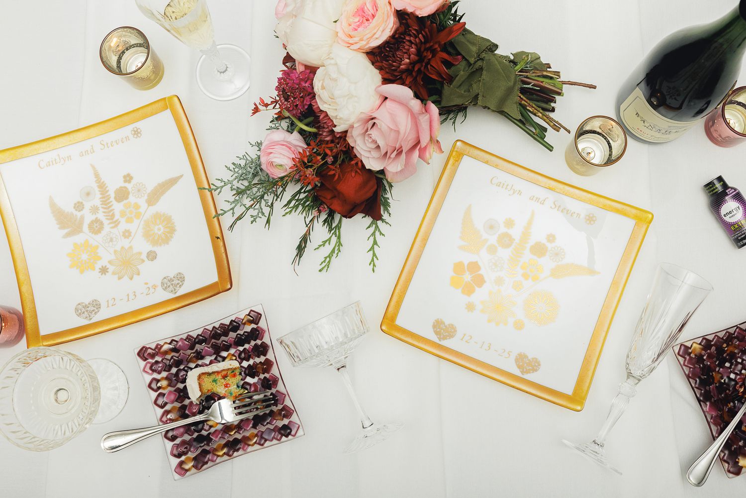 Wedding napkins with gold text alongside pink and burgundy flowers, rings, and wine on a white marble surface.