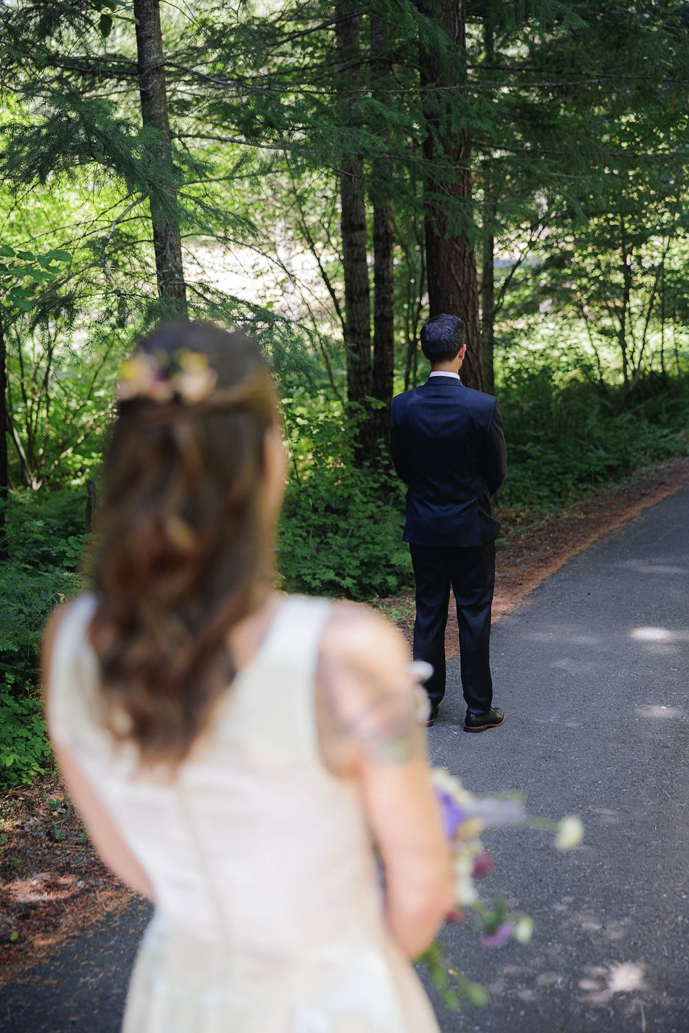 A bride approaches a figure in navy blue standing on a wooded path during a forest first look moment.