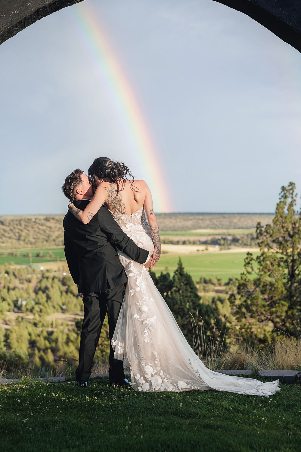 A couple embraces on a hilltop with a vibrant rainbow arching across the scenic landscape in the background.