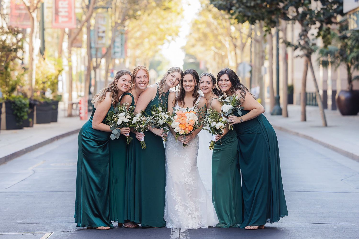 Wedding party with bridesmaids in emerald green dresses and bride in white posing together on a city street.