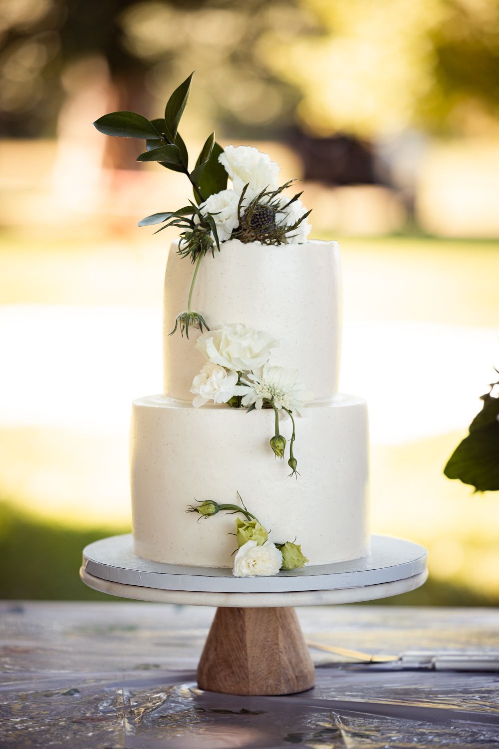 Elegant two-tier white wedding cake decorated with fresh greenery and flowers on a wooden cake stand.