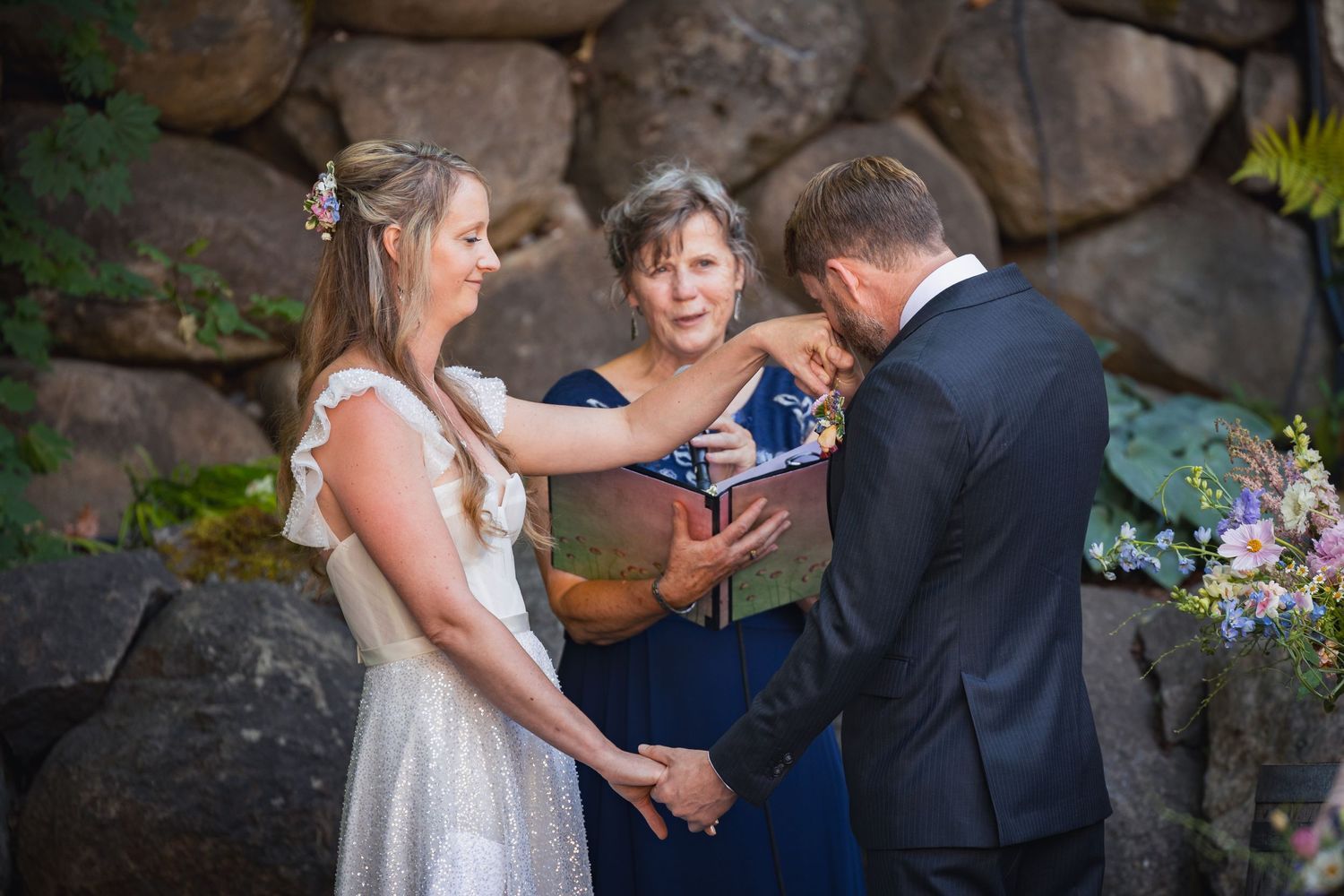 Wedding ceremony takes place outdoors against a stone backdrop with an officiant and couple holding hands.