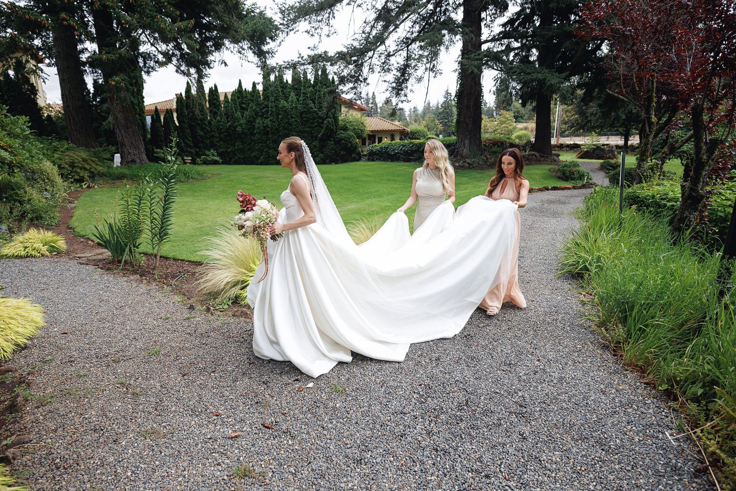 Bride in white gown walks garden path as two bridesmaids lift her flowing train at outdoor wedding venue.