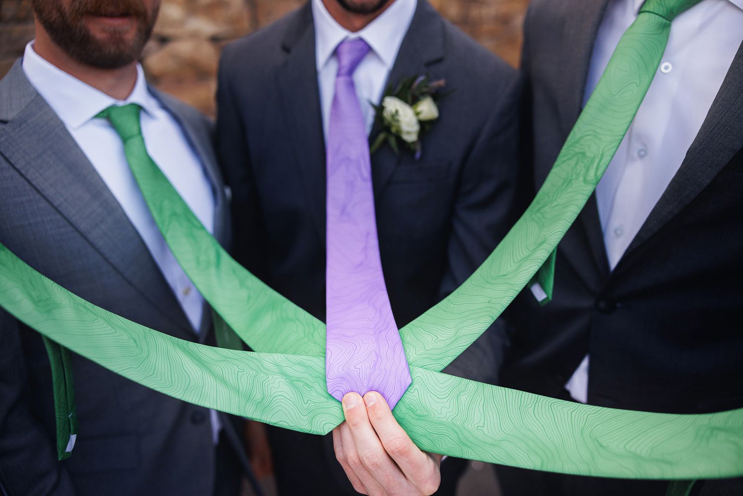 Three men in suits display green and purple ties, with one wearing a boutonniere, at a formal wedding event.