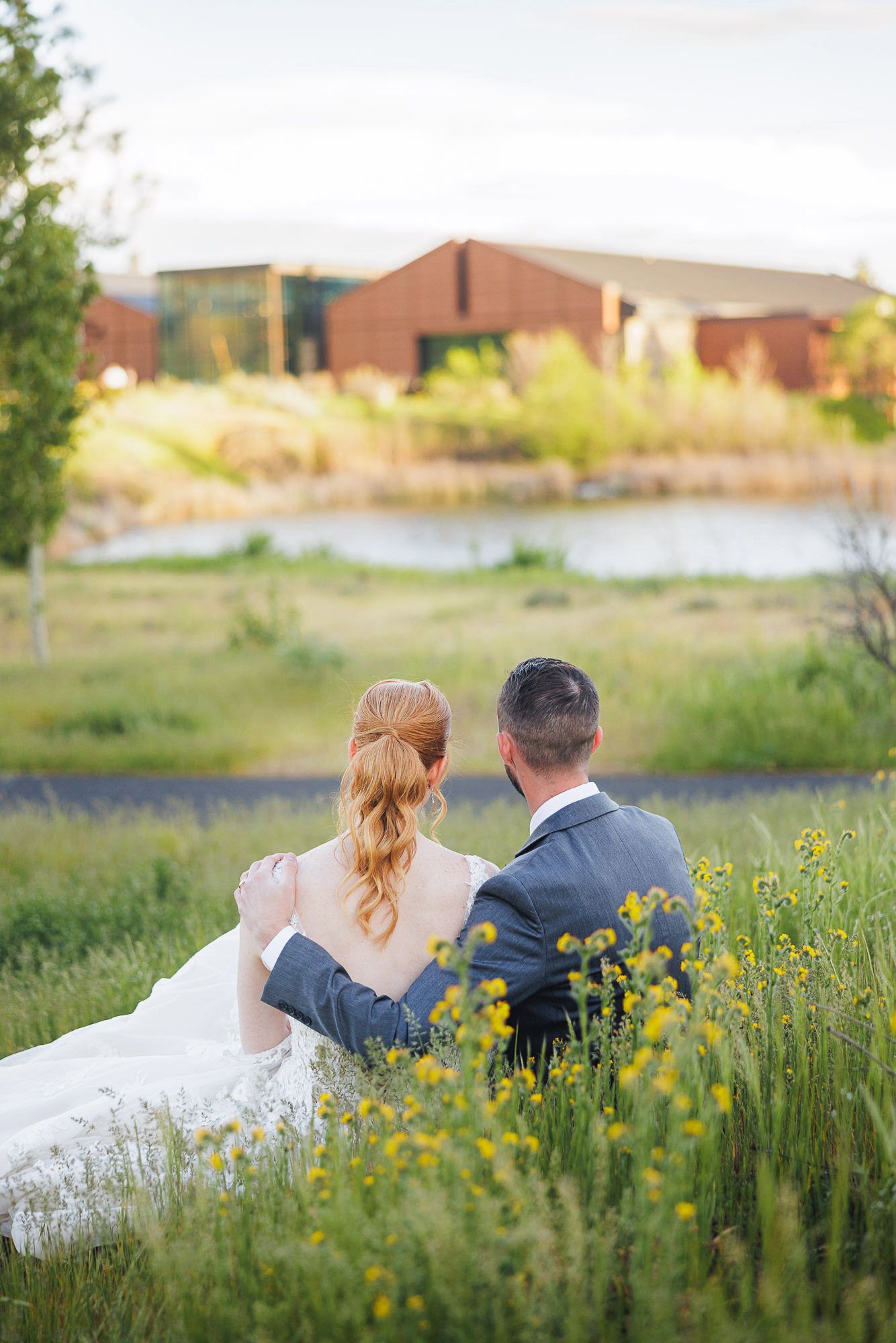 Bride and groom sitting together in a field of wildflowers, facing a serene pond and rustic building at golden hour.