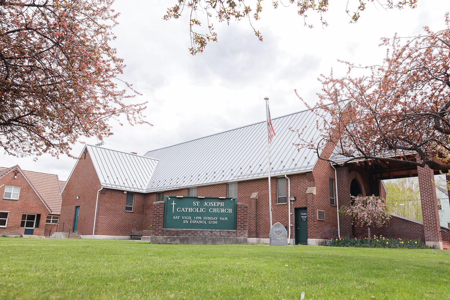St. Joseph Catholic Church brick building with metal roof, American flag, and green lawn surrounded by spring trees.