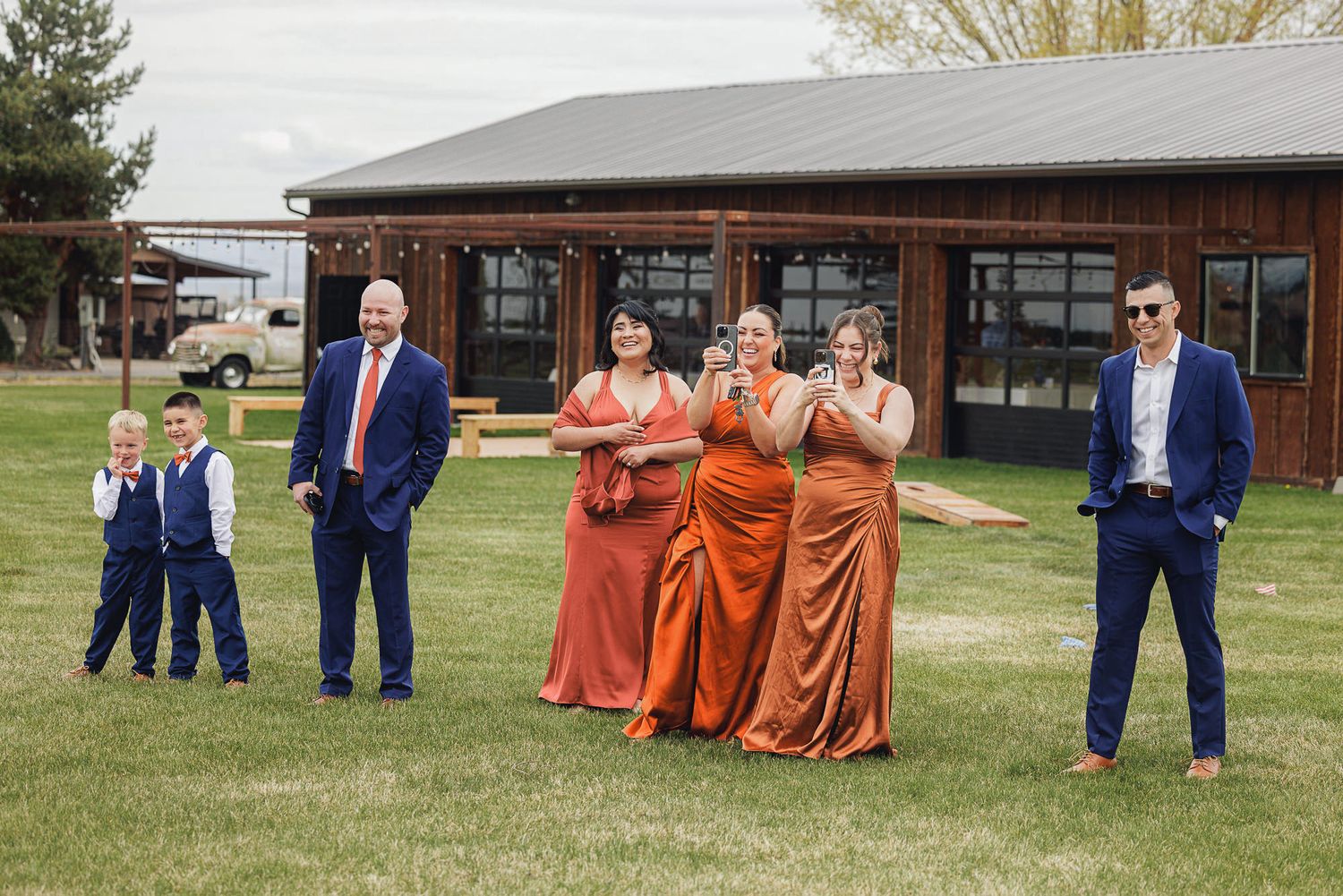 Wedding party in navy suits and orange satin gowns pose outdoors near a rustic barn venue on a sunny day.