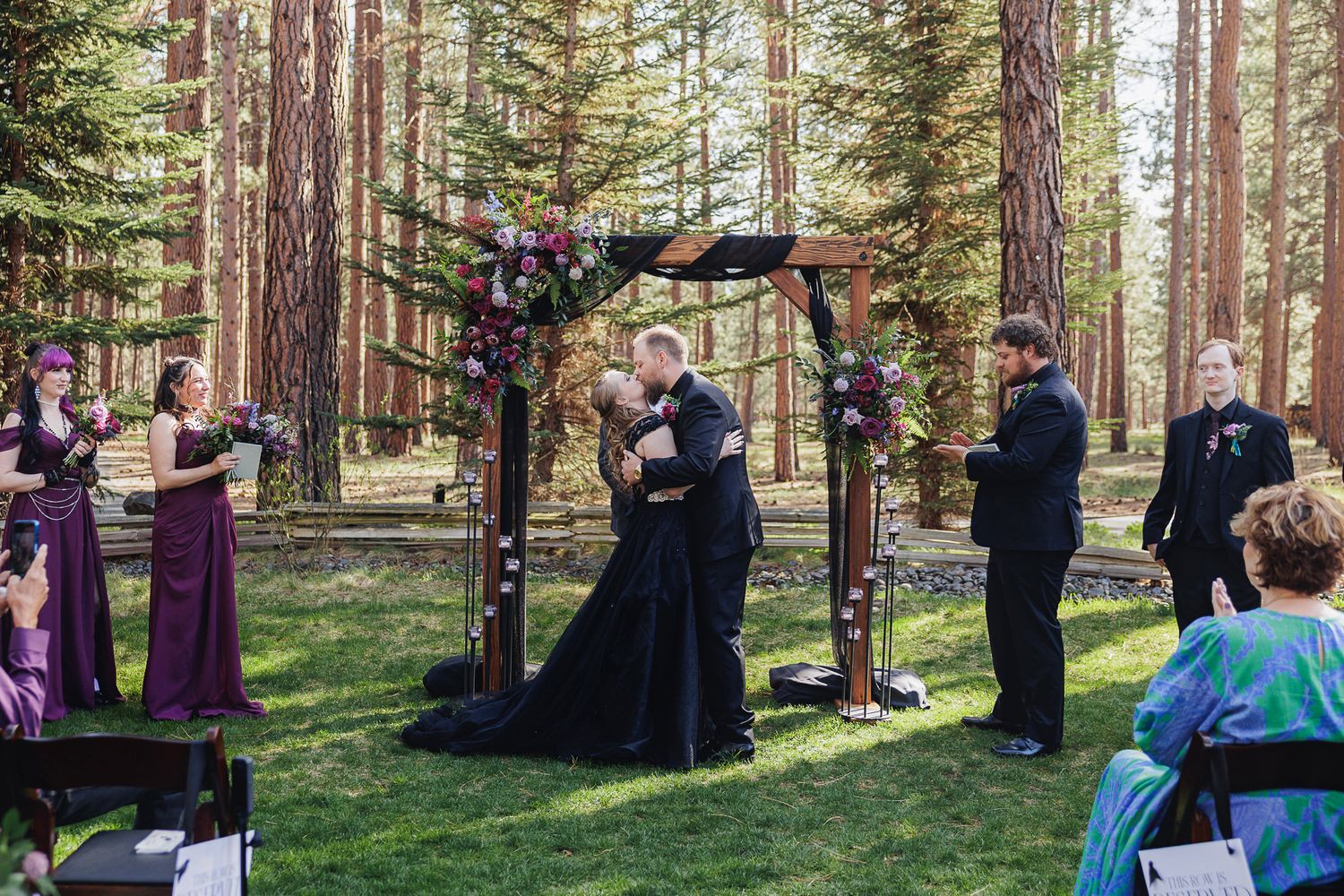 Couple sharing first kiss at outdoor forest wedding ceremony with purple bridesmaid dresses and floral arch.