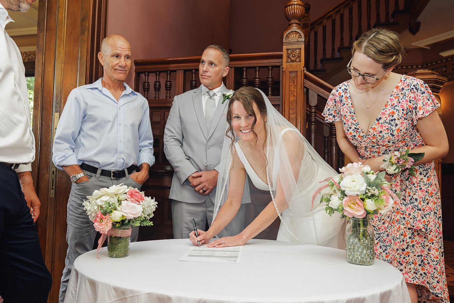 Wedding guests watching and signing the marriage certificate at an indoor ceremony with floral arrangements on the table.