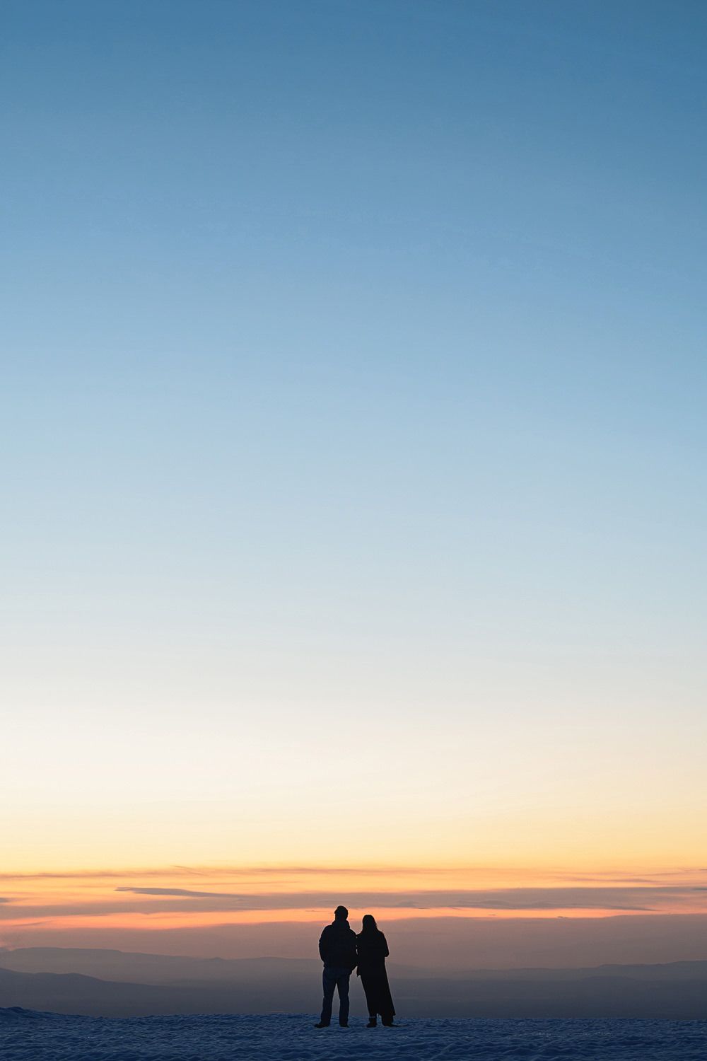Two silhouettes stand together against a colorful sunset sky on a snowy hilltop during winter.