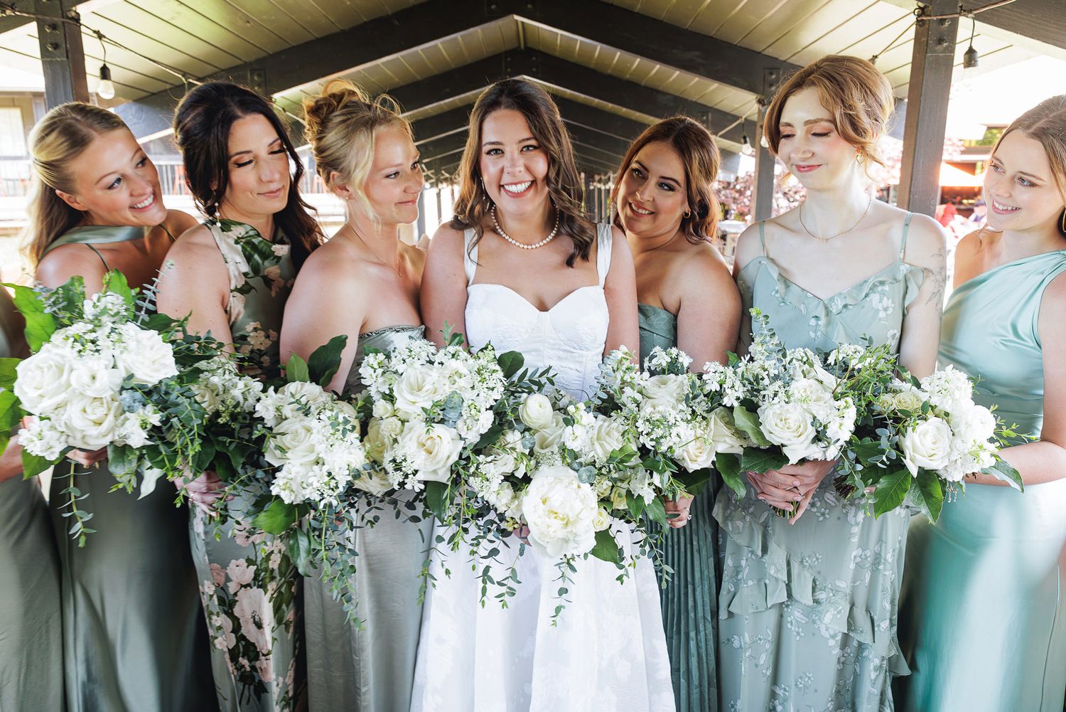 Bride in white gown with bridesmaids in sage green dresses holding white floral bouquets with greenery at wedding.