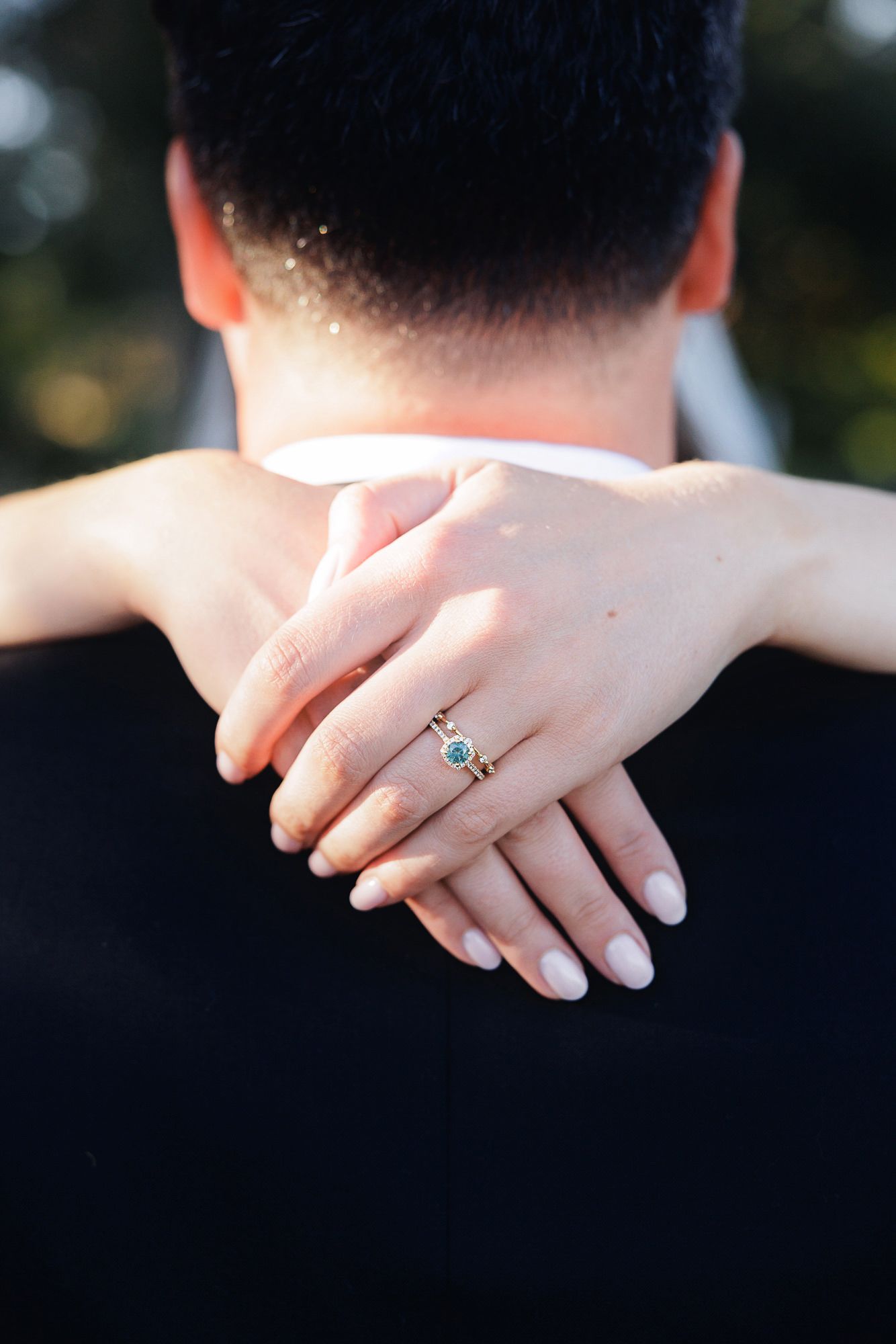 Close-up of a woman's hand with an emerald engagement ring resting on a man's shoulder during a romantic embrace.