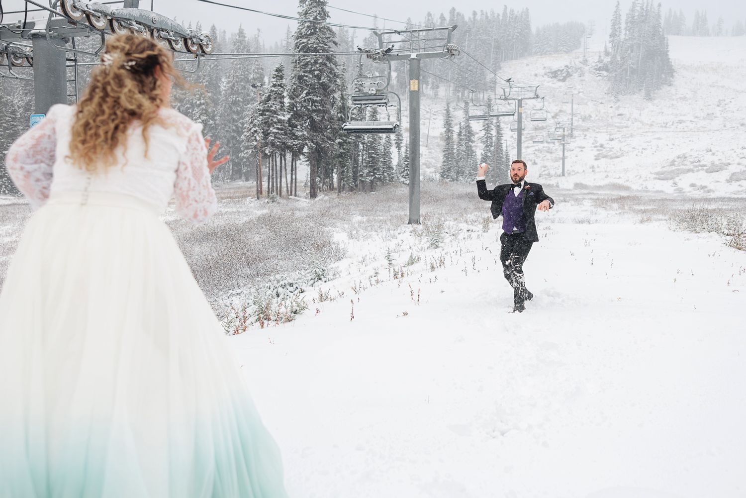 A winter wedding couple walks through snowy mountains with ski lifts and snow-covered trees in the background.