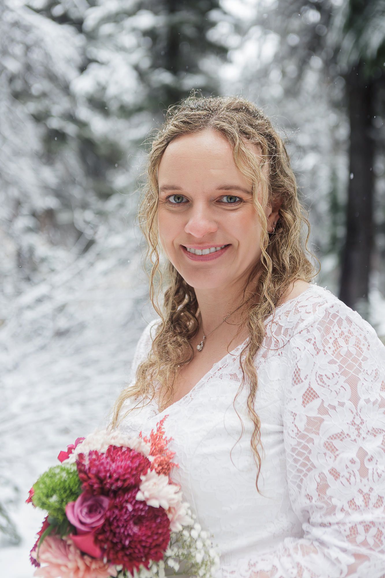 A bride in a white lace dress holds a vibrant bouquet of pink and burgundy flowers against a snowy winter backdrop.