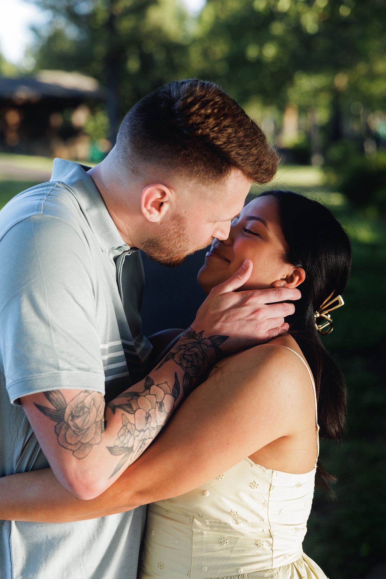 A tattooed man tenderly holds a woman's face while they share a romantic kiss outdoors in a sunlit green park.