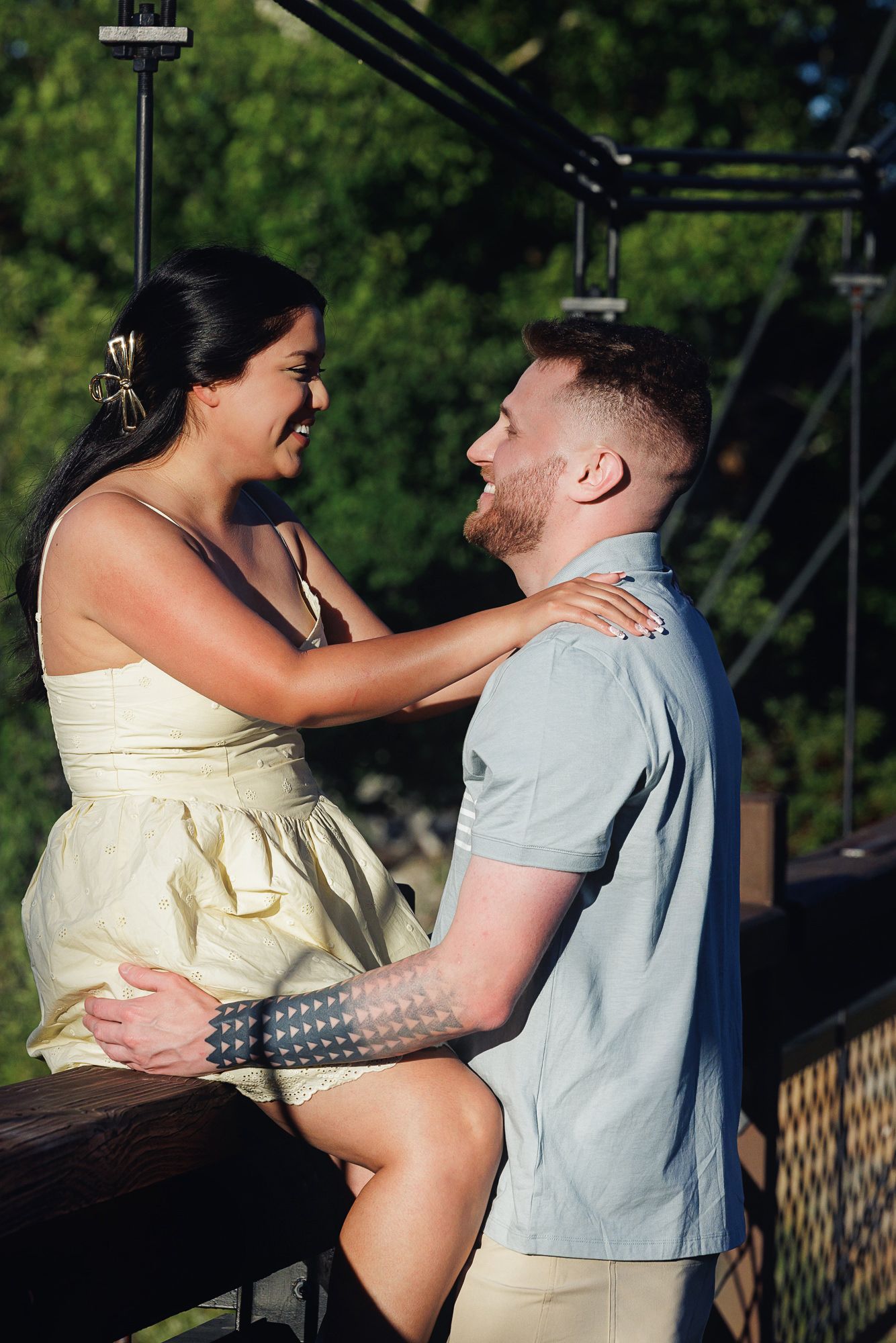 A smiling couple sharing a romantic moment outdoors, woman in yellow dress sitting on railing with tattooed man in gray shirt.