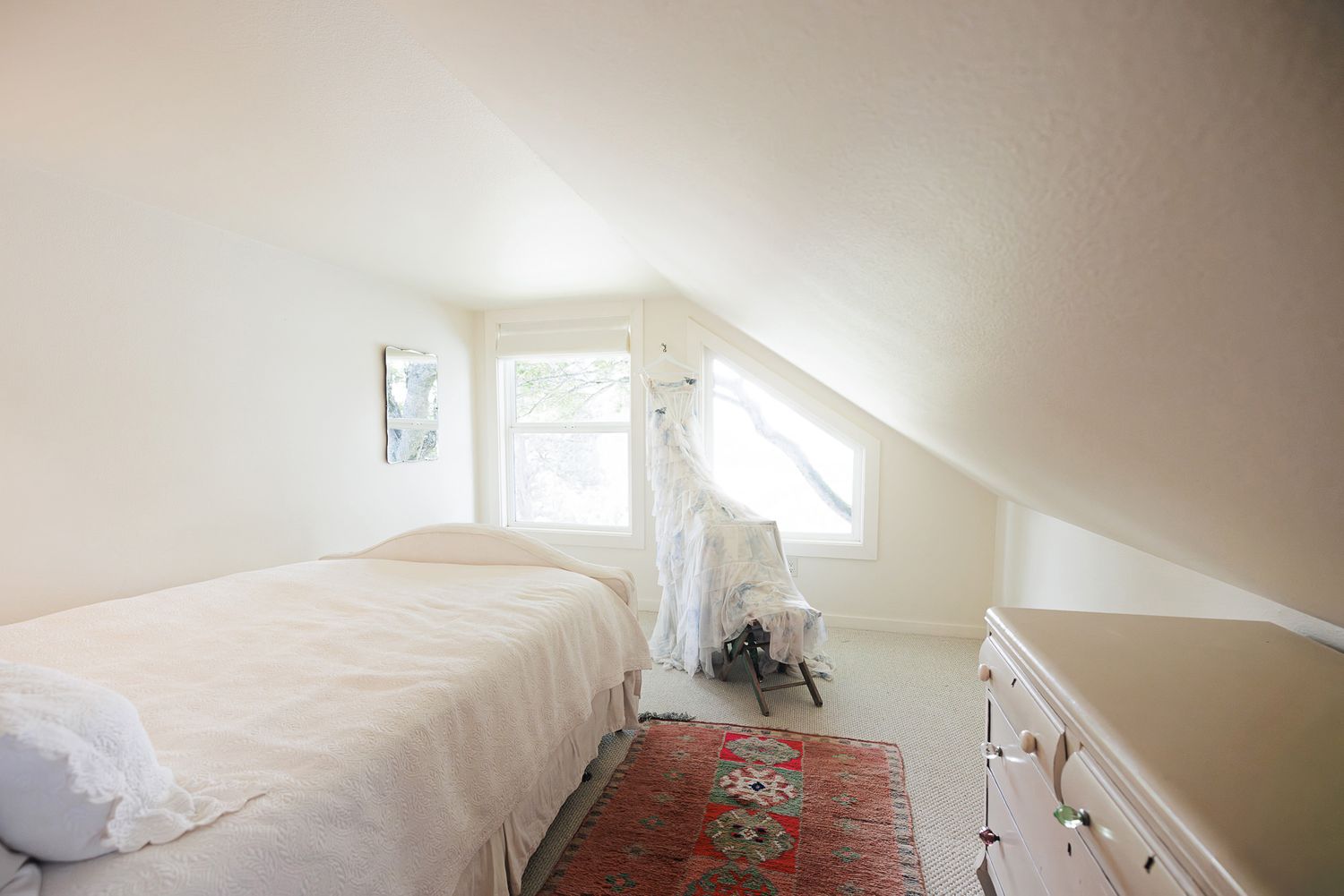 Bright attic bedroom with sloped ceiling, white bedding, hardwood floors, red rug, and natural light from two windows.