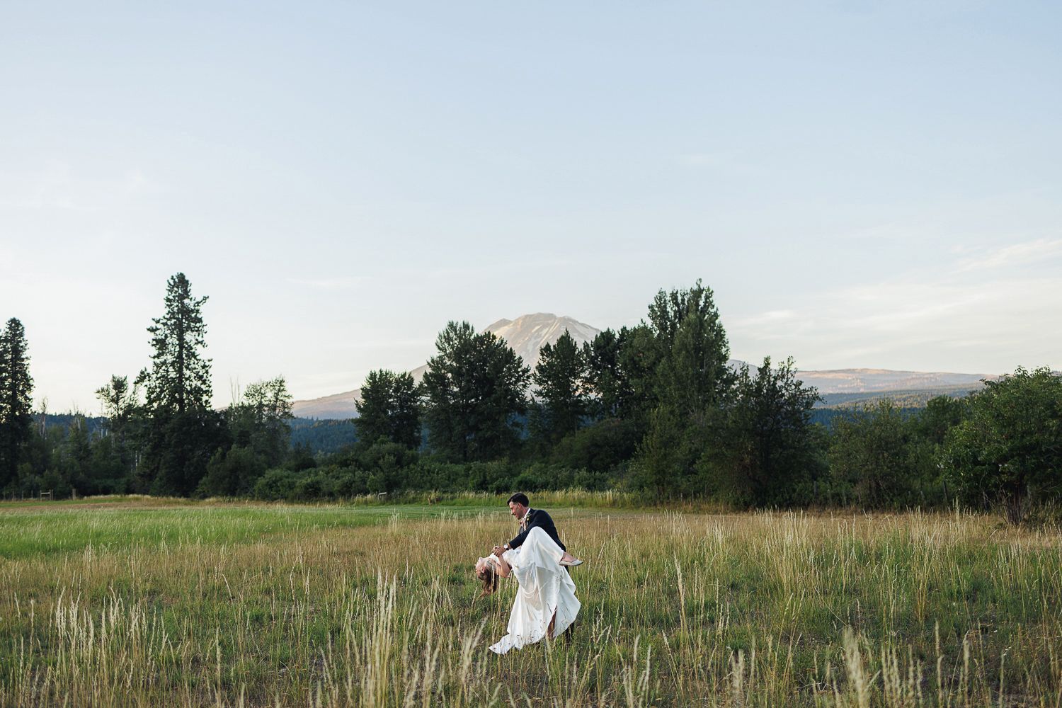 A person in a flowing white dress walks through a serene meadow with trees and mountains in the background at dusk.