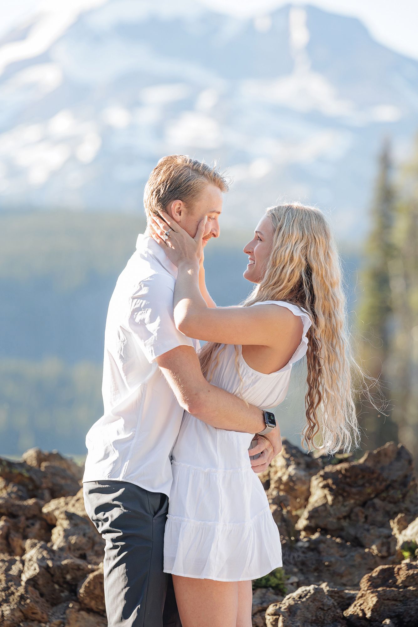 A couple embraces and smiles at each other on rocky terrain with snow-capped mountains and pine trees in the background.