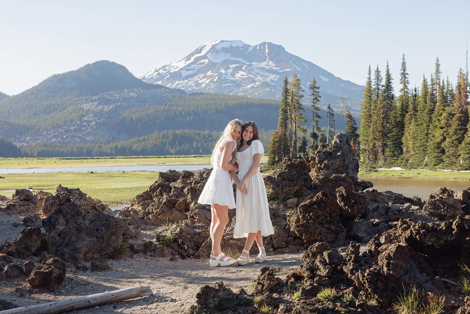 Two women in white dresses embrace among lava rocks with a snow-capped mountain and pine trees in the background.
