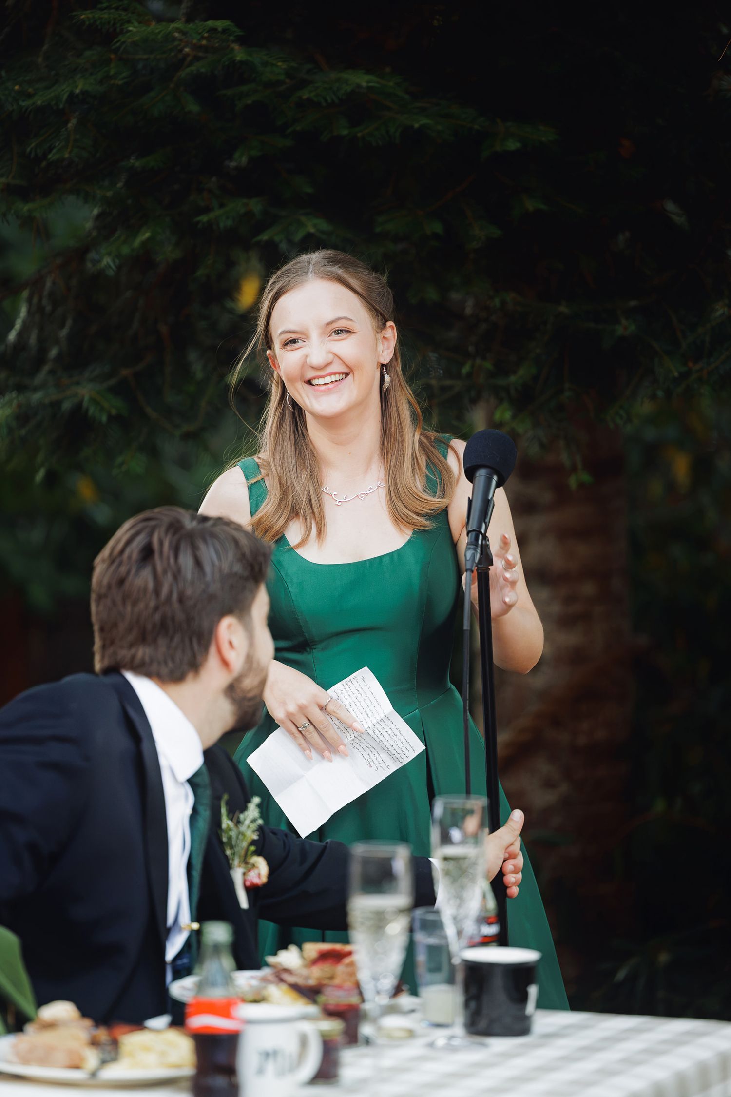 A smiling woman in a green dress gives a speech at a wedding reception, holding notes at a microphone outdoors.
