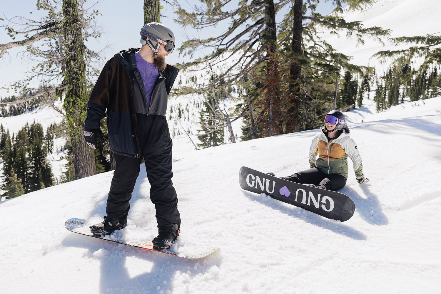 Two people snowboarding on a snowy mountain slope surrounded by pine trees.