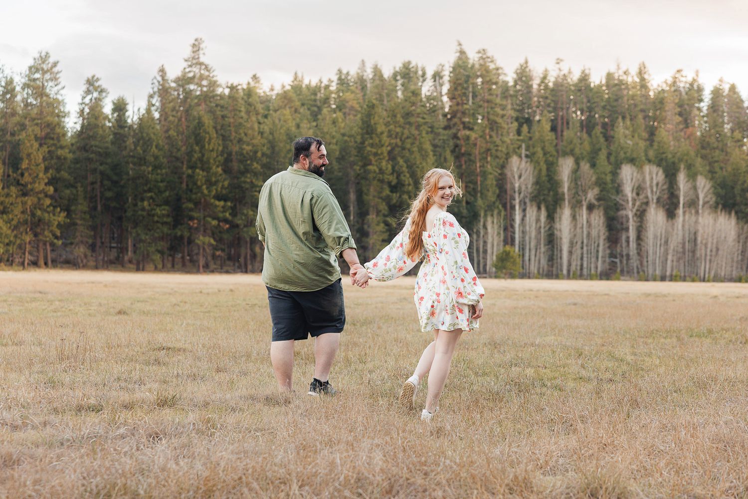 Couple holding hands walking through a meadow with tall pine trees in the background.