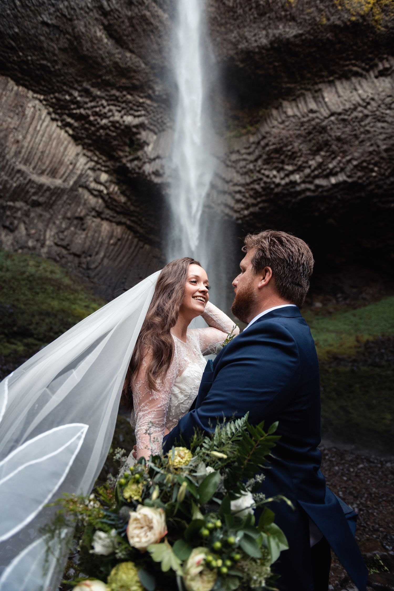 Couple in wedding attire poses near a majestic waterfall while holding an elegant bouquet of white and green flowers.