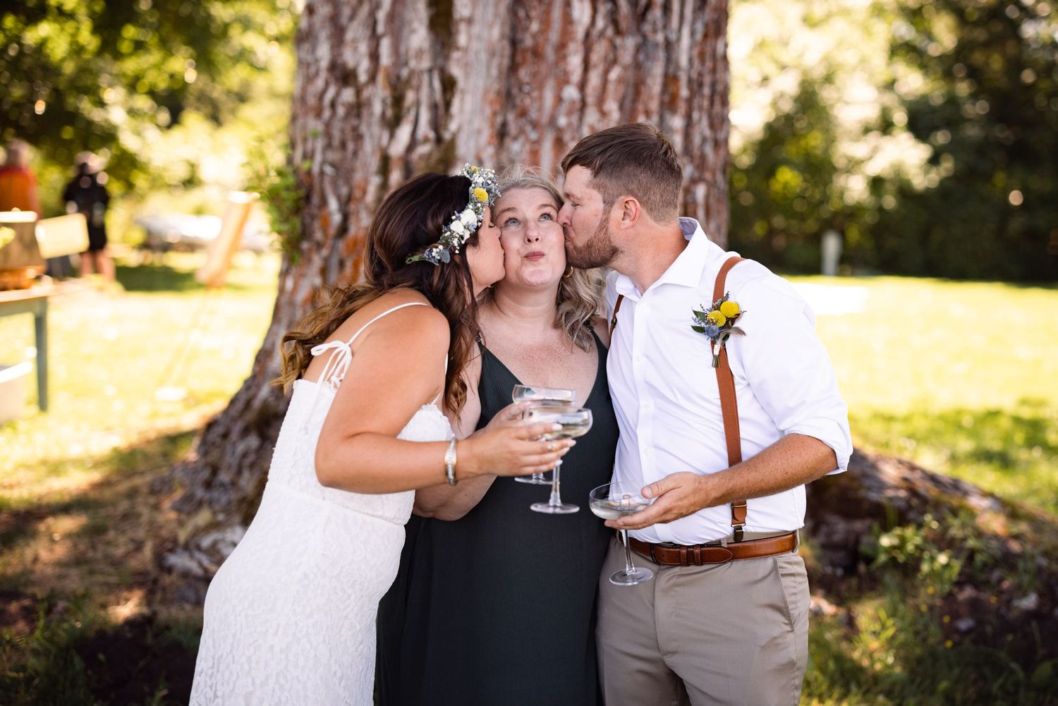 Three people share a celebratory drink together outdoors at a wedding reception near a large tree trunk.